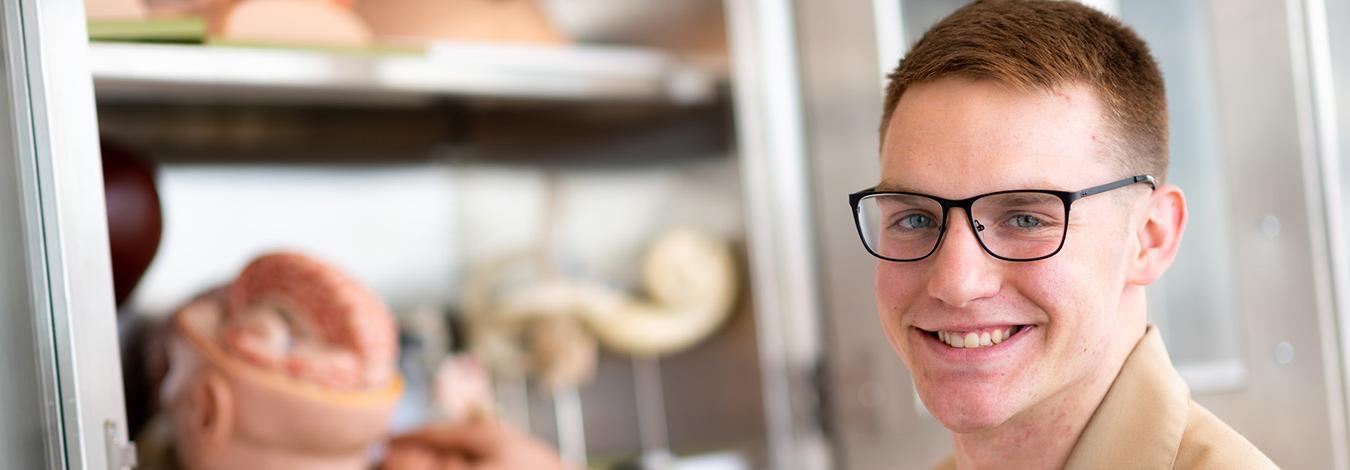 Young man wearing glasses, military dress shirt, with one hand in a cupboard of medical anatomy models.