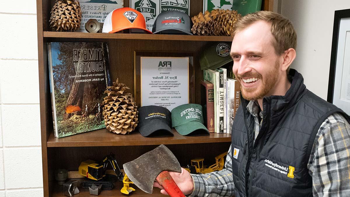 Smiling man holds axe in front of bookshelf of logging memorabilia.