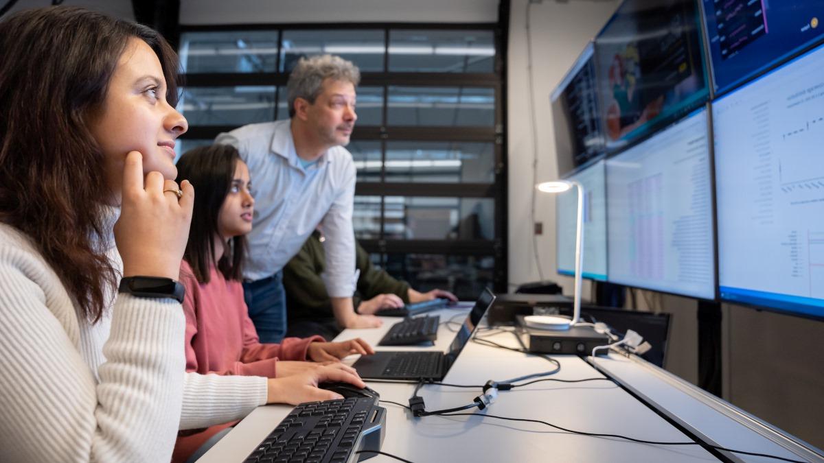 Two students with a professor looking over monitors in a Cybersecurity class.